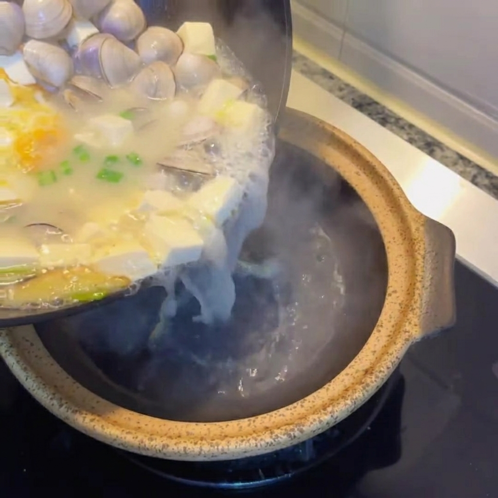 Tofu and clam soup being poured into a rustic clay serving pot.
