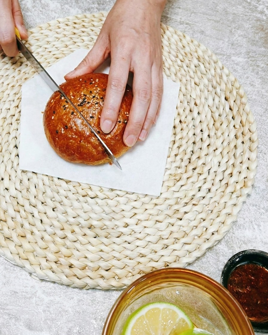 A close-up of a hand using a knife to slice a soft burger bun on a woven mat.