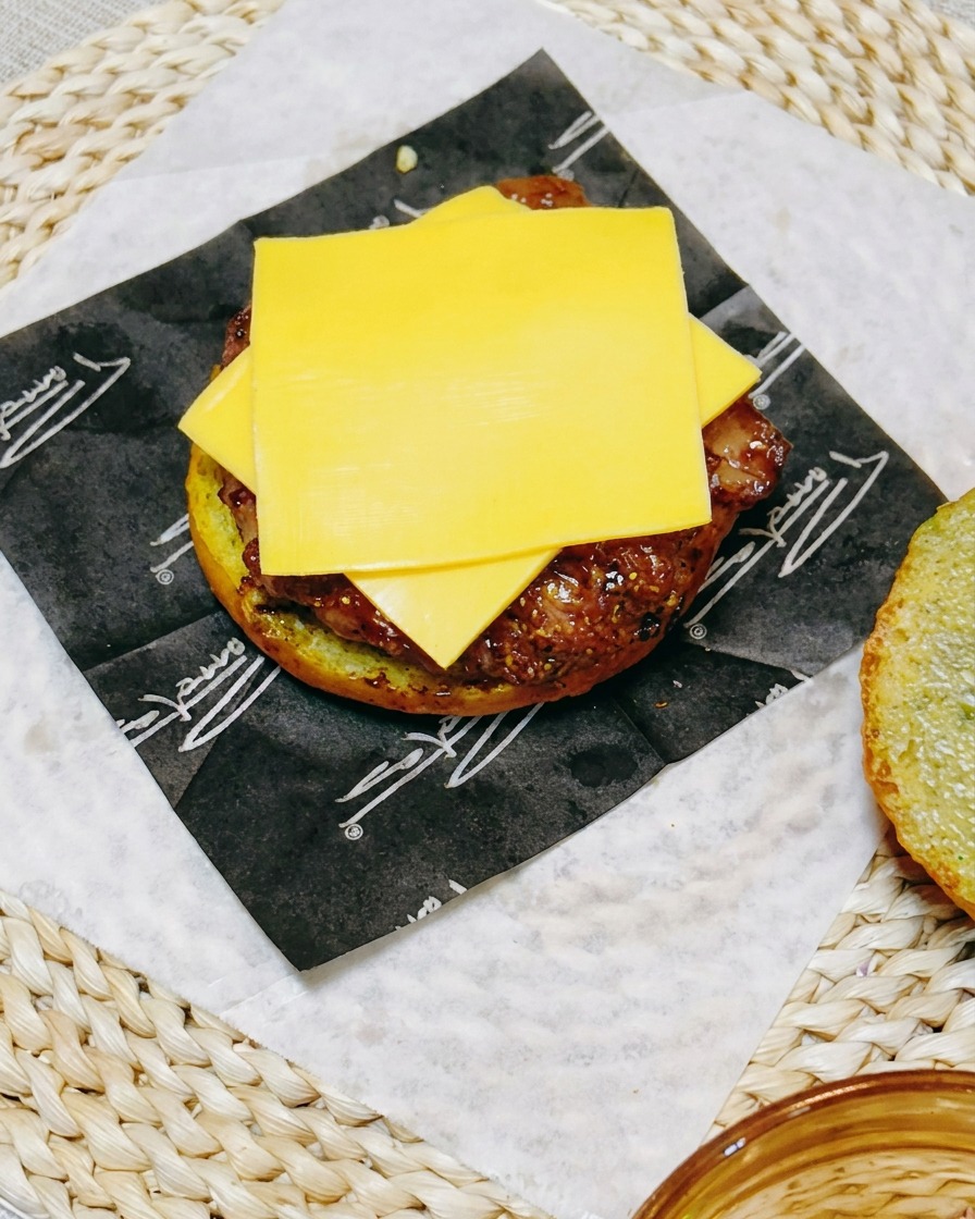 A close-up of a burger assembly showing a slice of processed cheese resting on the chicken patty, topped with fresh cucumber rounds.