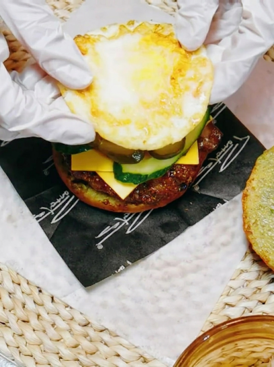 A pan-fried teriyaki chicken thigh being placed on top of tomato and lettuce on a bun.