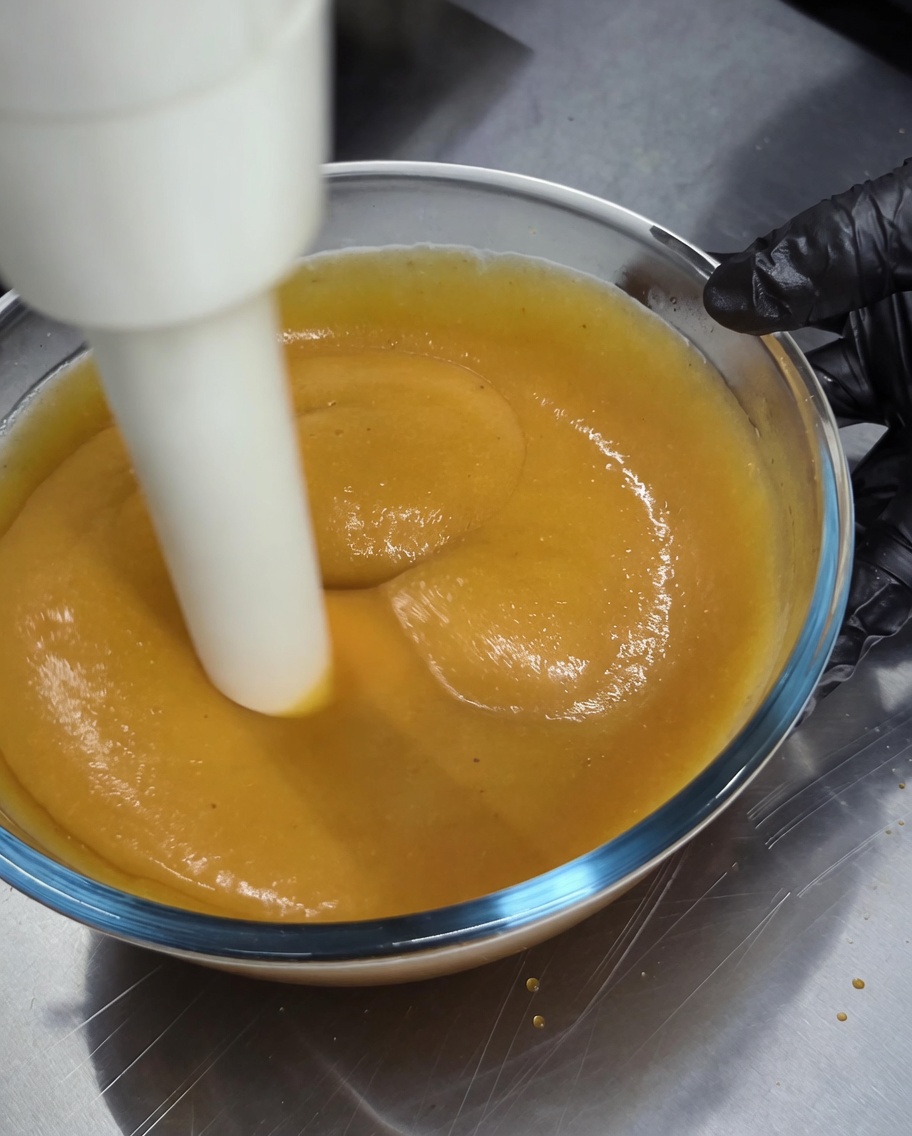 An immersion blender being used to blend a thick, golden-brown sauce in a glass bowl.
