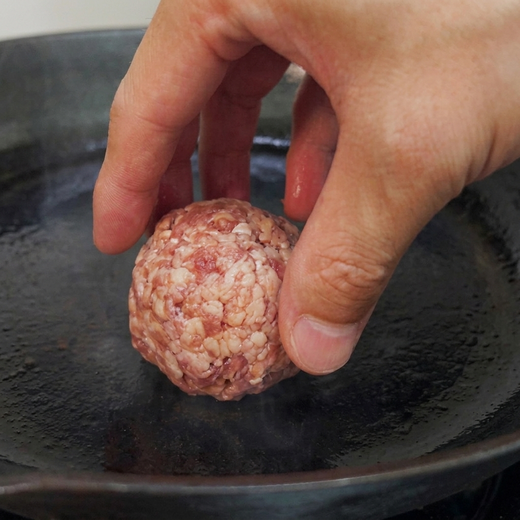 Raw beef balls being placed into a preheated flat-bottomed pan, ready for searing.