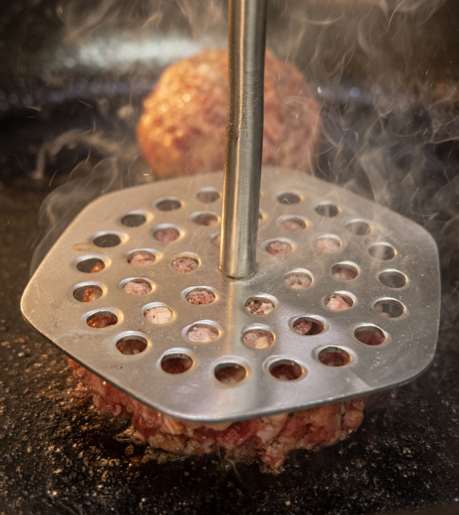 A metal meat press being used to flatten beef balls into thin burger patties inside a hot pan.