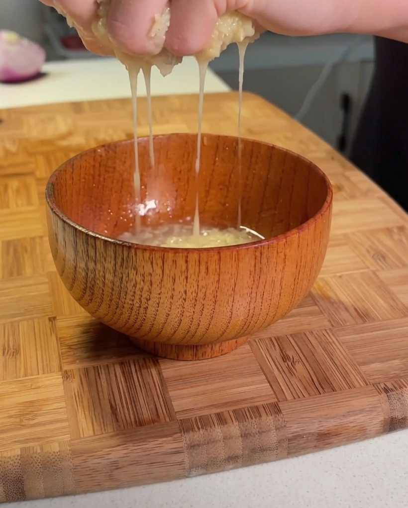 Hands squeezing shredded potato over a small wooden bowl to extract liquid.