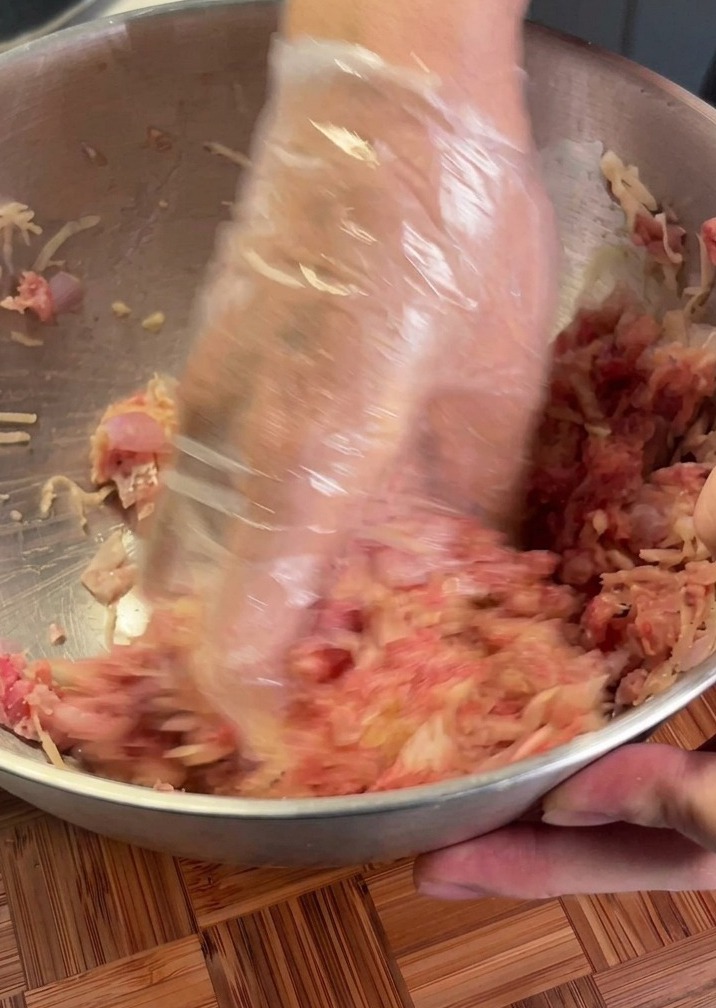 Hands in a plastic glove mixing raw beef and shredded potato in a metal bowl.
