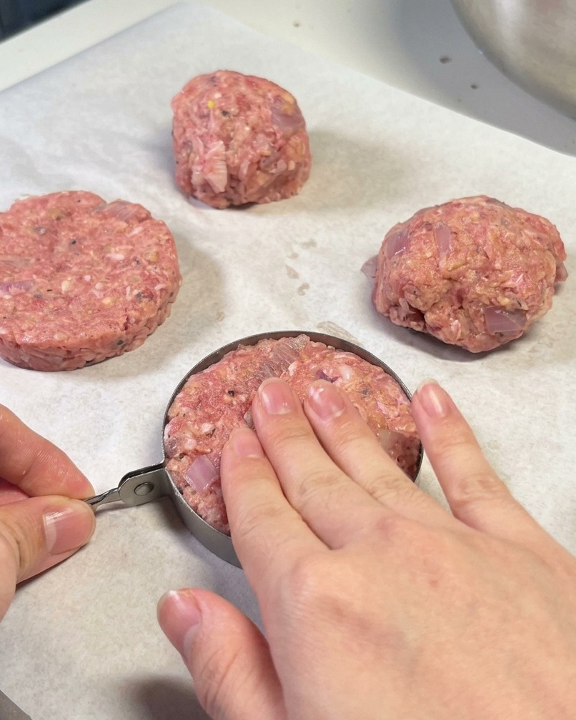 Hands using a metal ring mold to shape a hamburger patty on parchment paper.