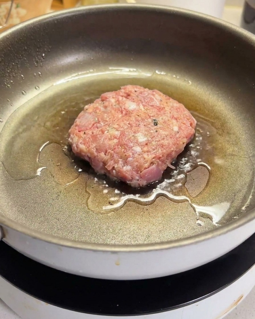 A raw ground beef patty sizzling in a hot, oiled non-stick pan.