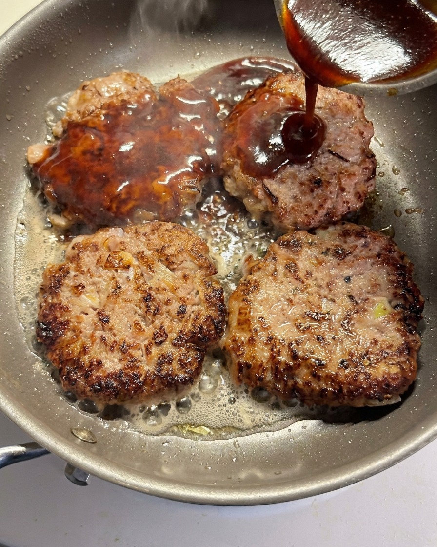 The sauce being poured over four browning beef patties in a frying pan.
