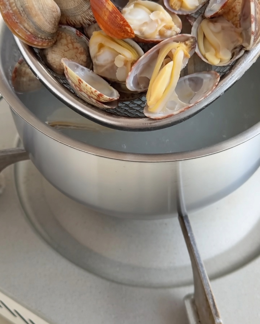 Freshly cooked clams in shells held over a pot, with clam meat being carefully removed.