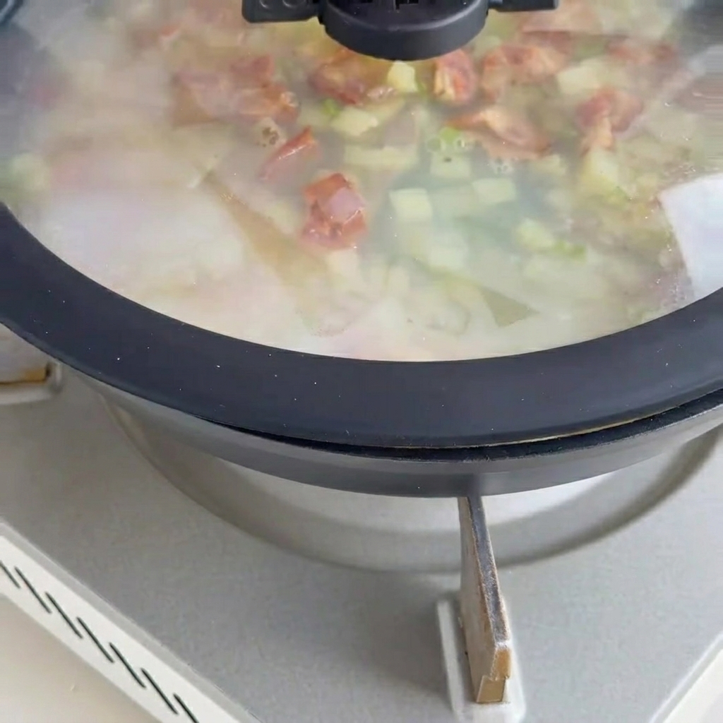 Creamy clam chowder base simmering in a pan with tender clam meat and visible bacon bits on top.
