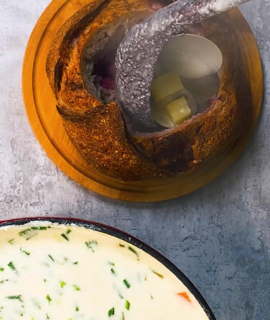 Rich, creamy clam chowder being poured from a ladle into a rustic bread bowl, garnished with golden-brown crispy bacon pieces.