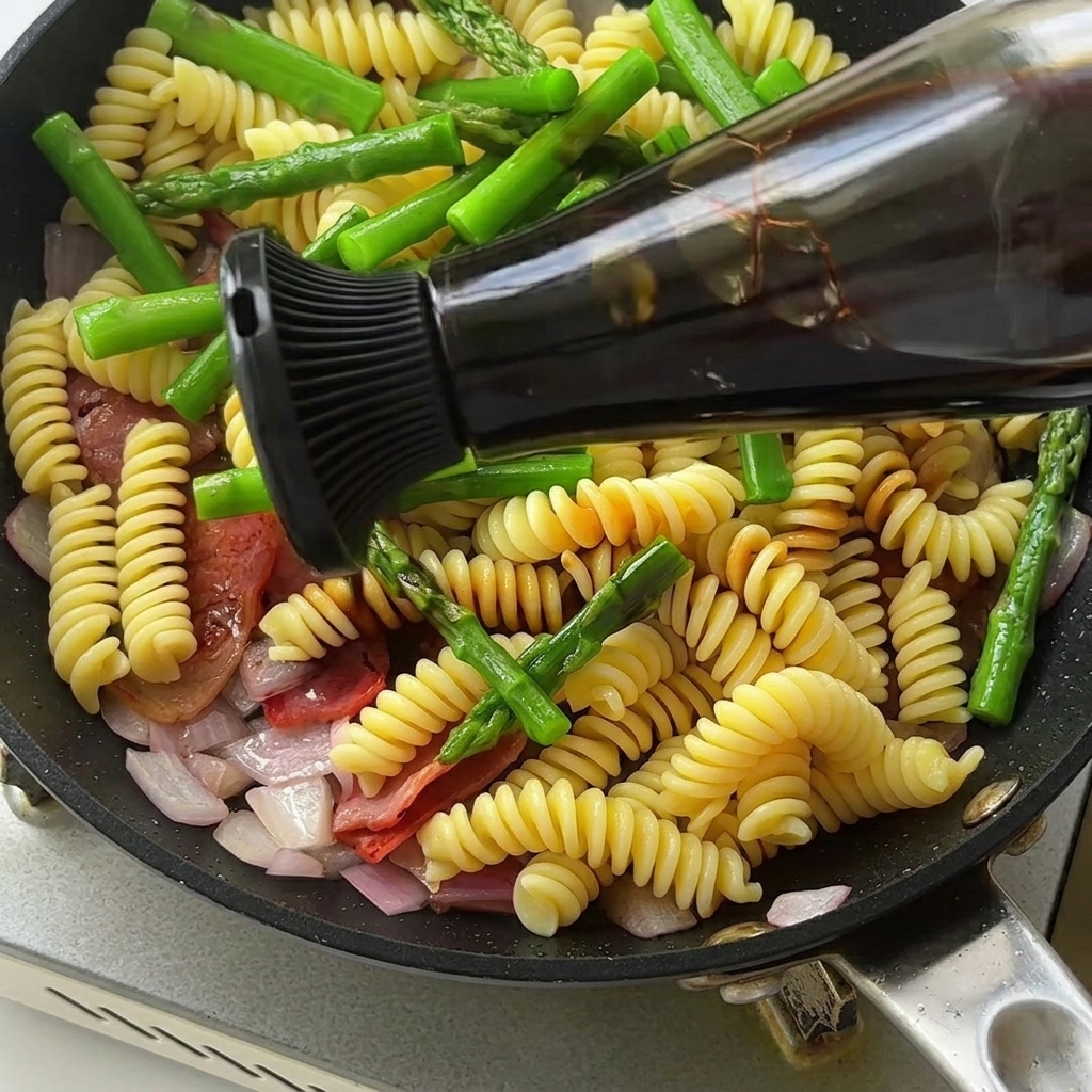 A close-up of a pan containing fusilli pasta, sautéed asparagus, and bacon being seasoned with a dark, rich sauce.