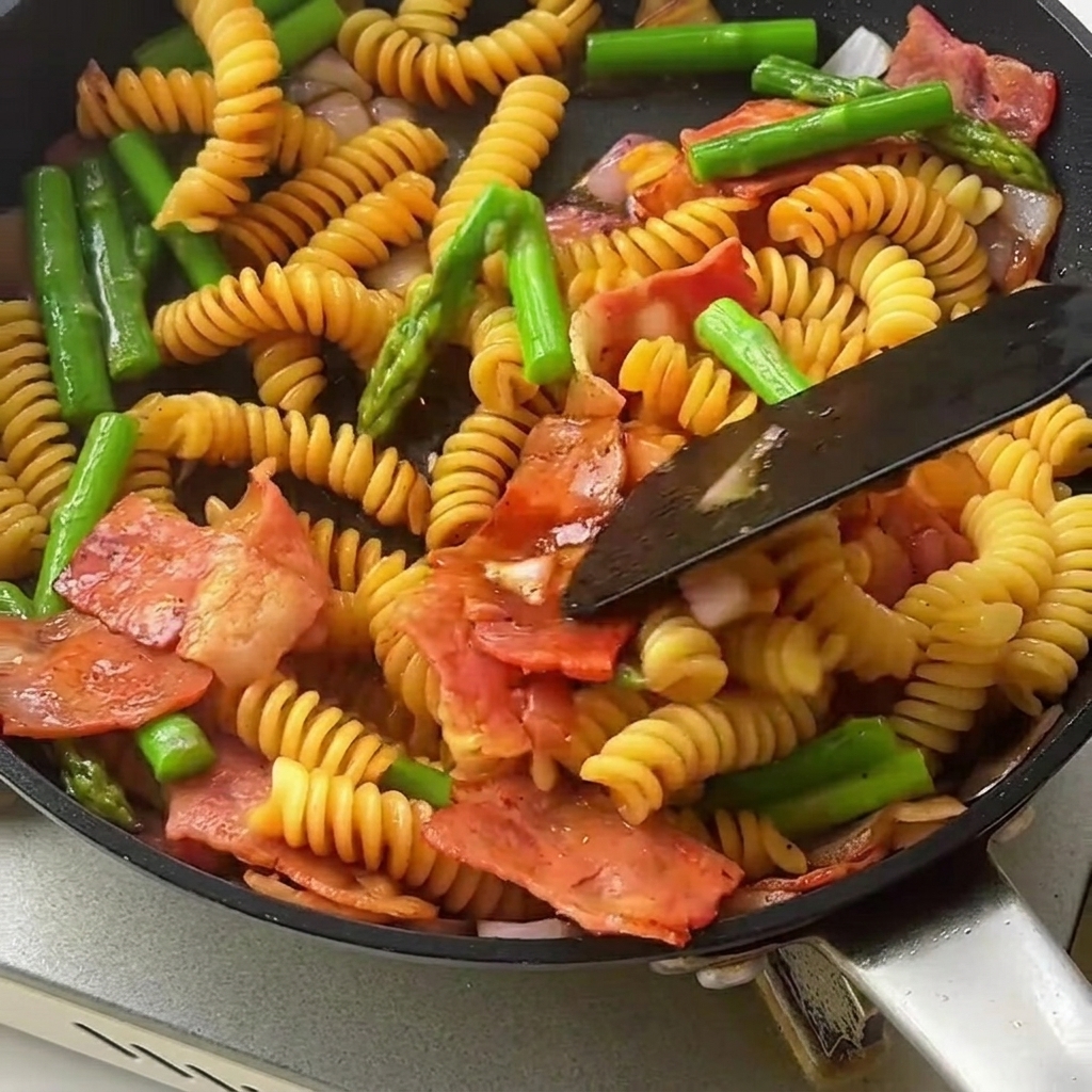 A spatula tossing together the saucy, glazed fusilli pasta, crisp asparagus, and savory bacon pieces in a large skillet.