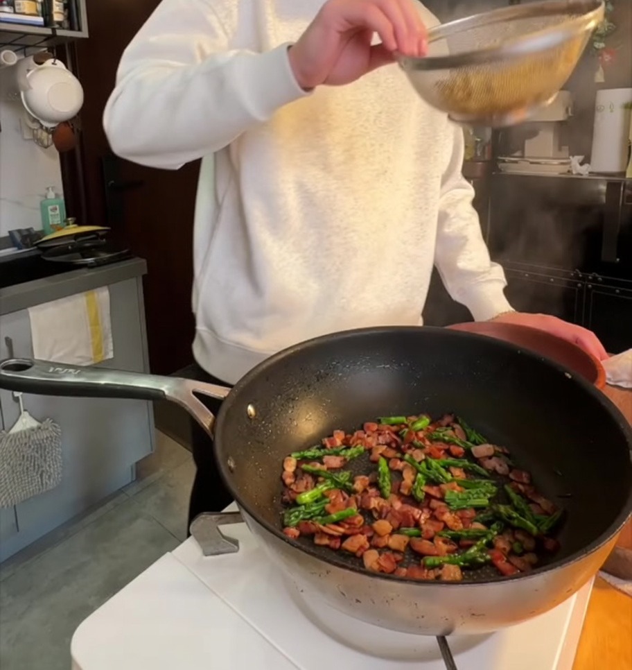 Boiled penne pasta being added to a pan with crispy bacon and sautéed asparagus tops.