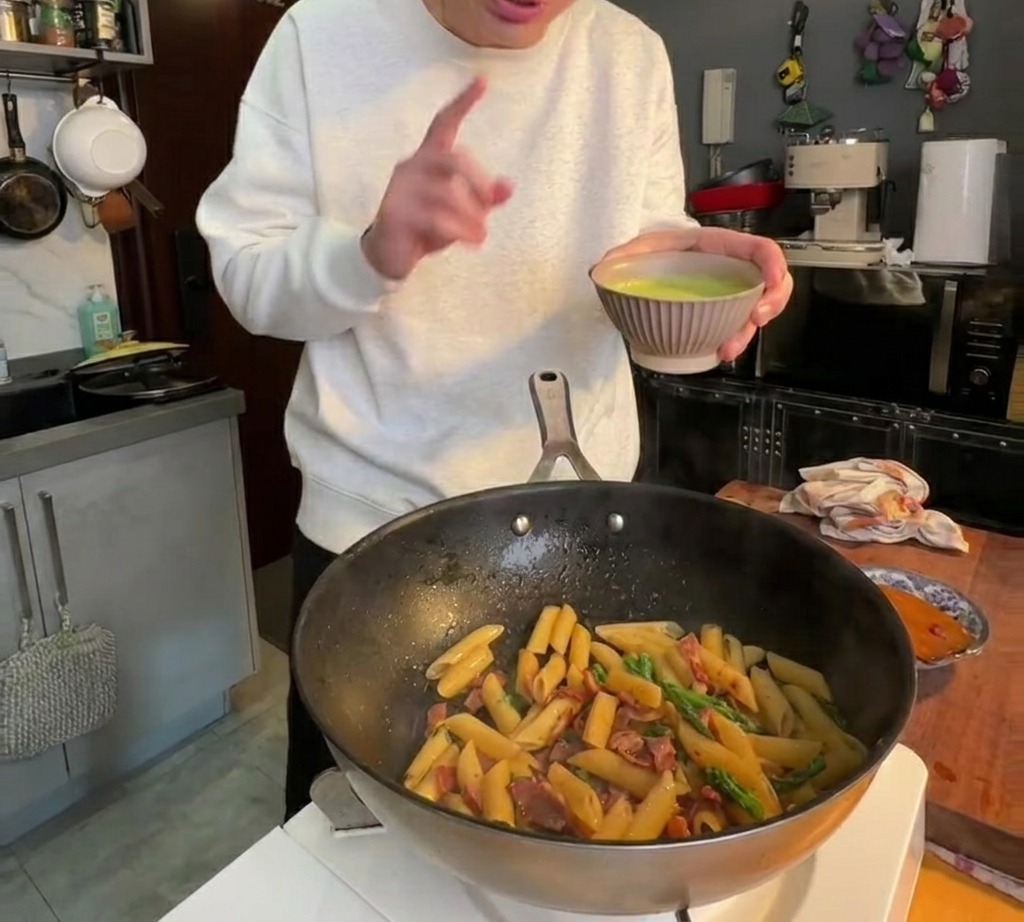 Smooth green asparagus sauce being poured over penne pasta in a pan and stirred to coat.