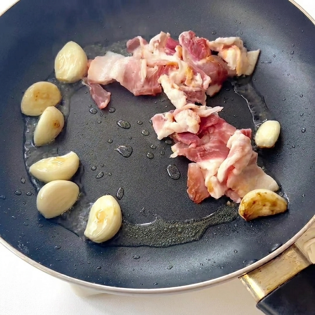Bacon frying in a pan alongside browned garlic cloves.