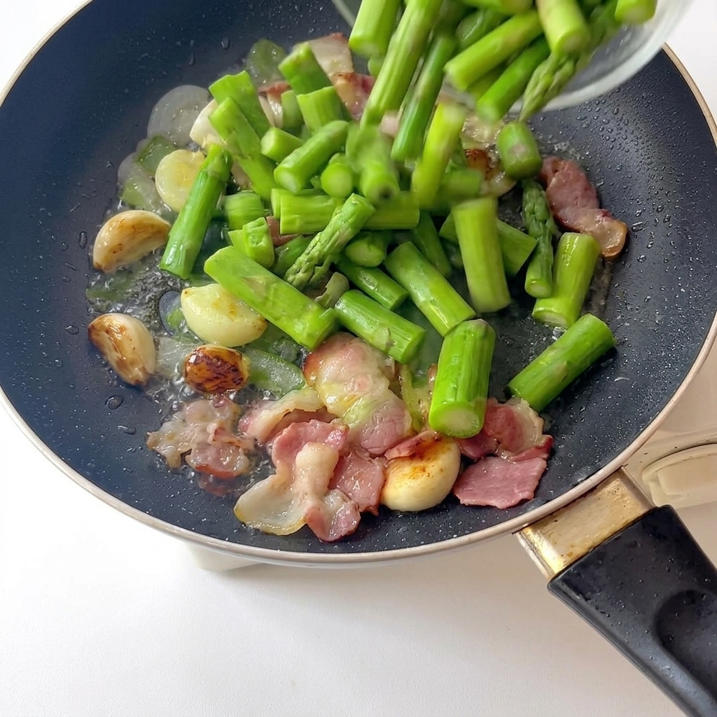 Freshly blanched bright green asparagus pieces being poured into a pan containing fried garlic and browned bacon strips.