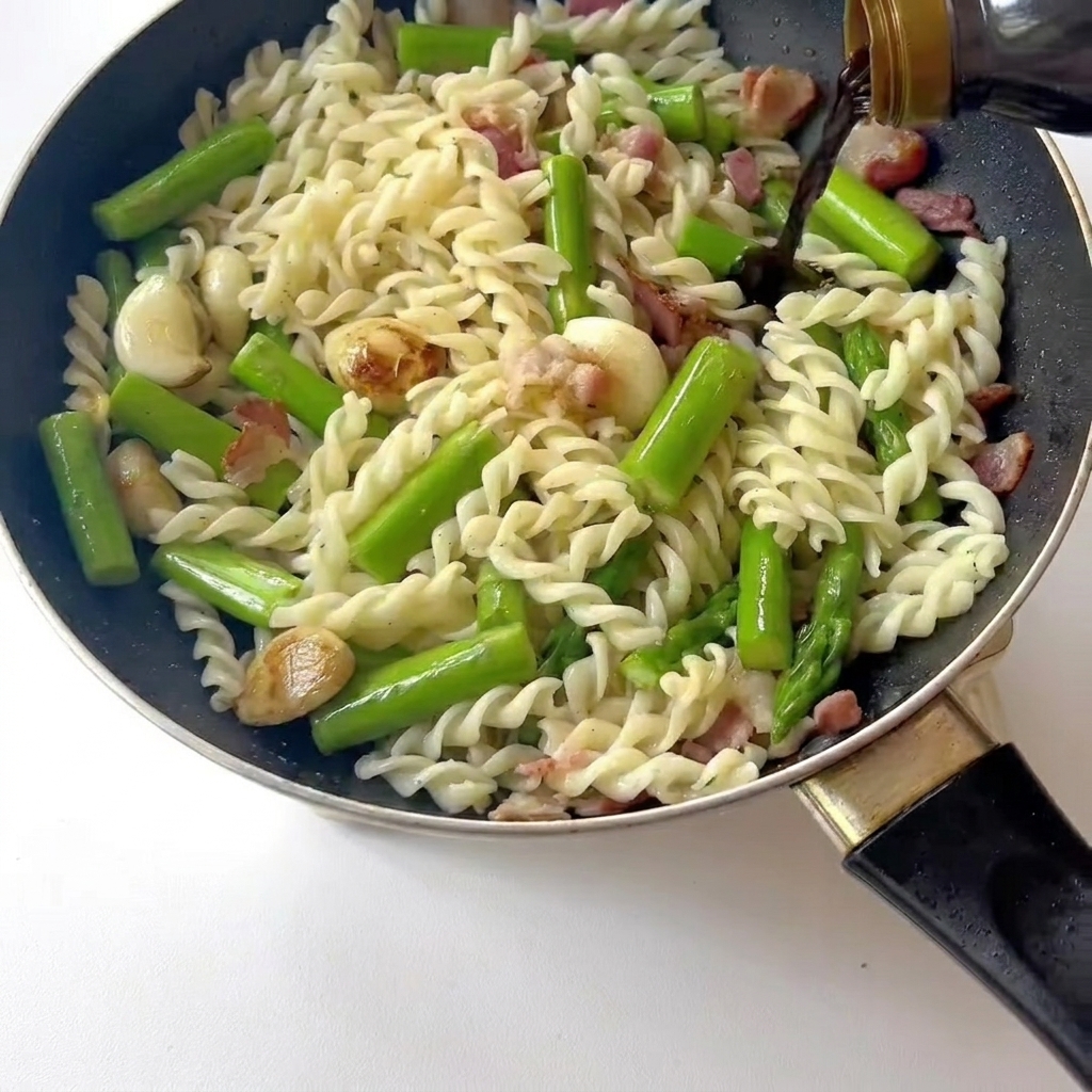 Close-up of a spatula stirring pasta, bacon, and asparagus in a pan as light soy sauce is being poured over it.