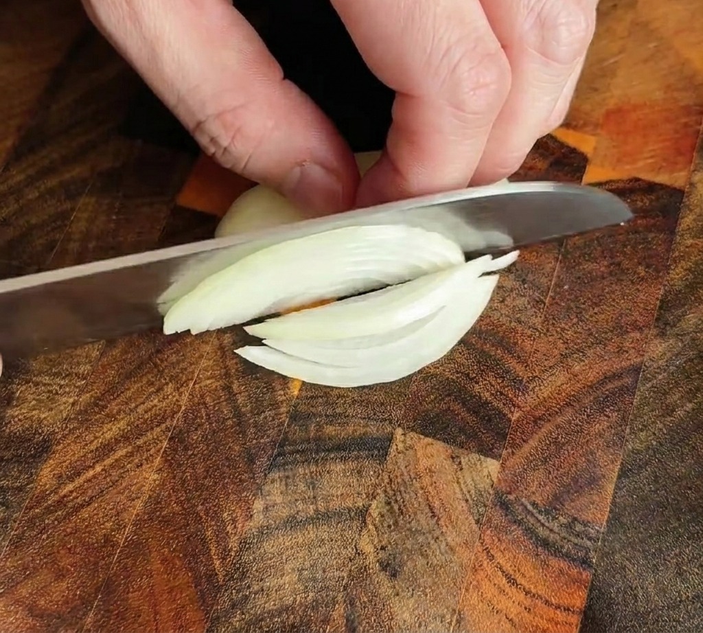 Chefs knife slicing an onion into thin strips on a wooden board.