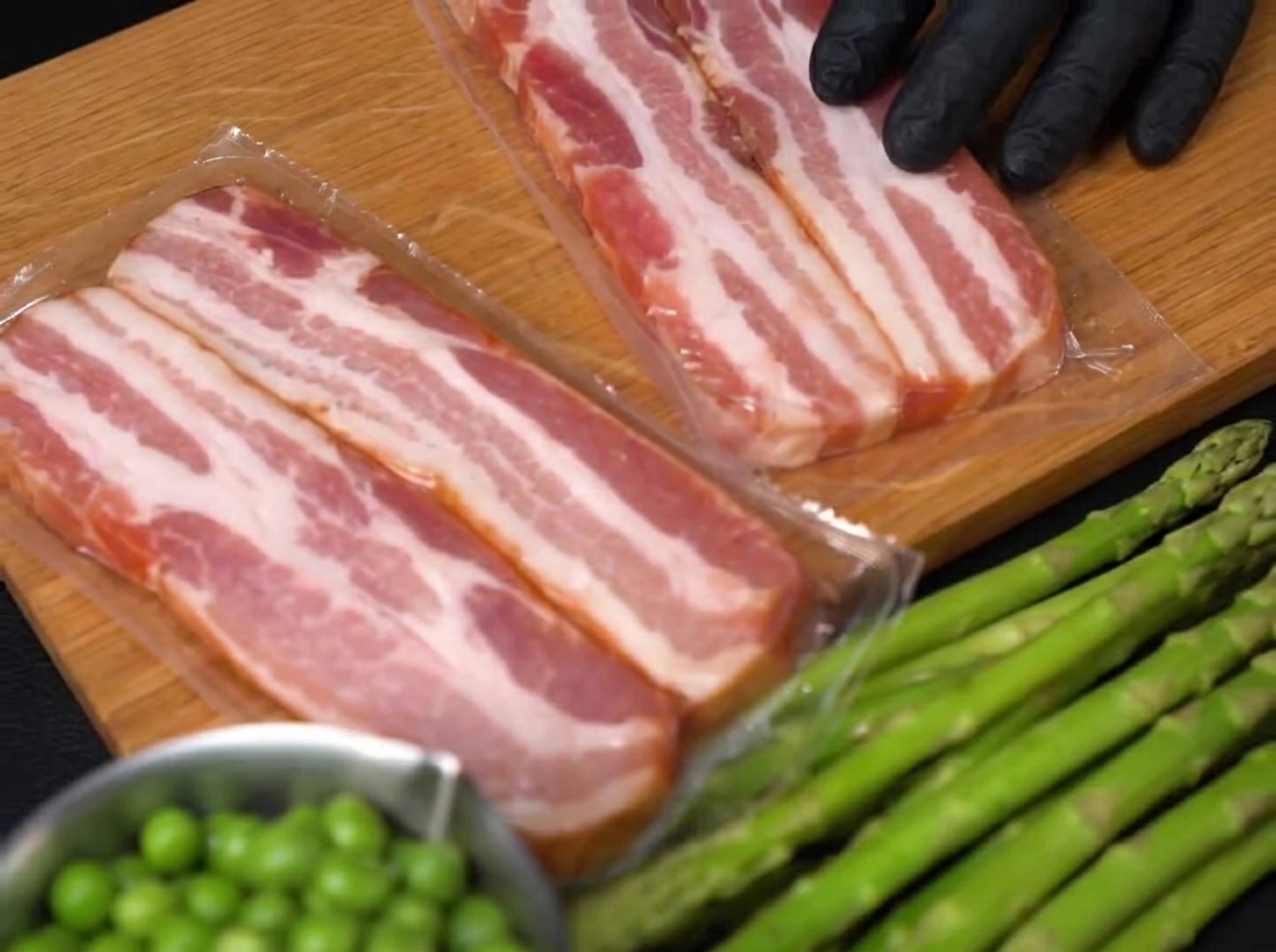 A close-up view of a gloved hand using a knife to slice thick strips of raw bacon on a wooden board.
