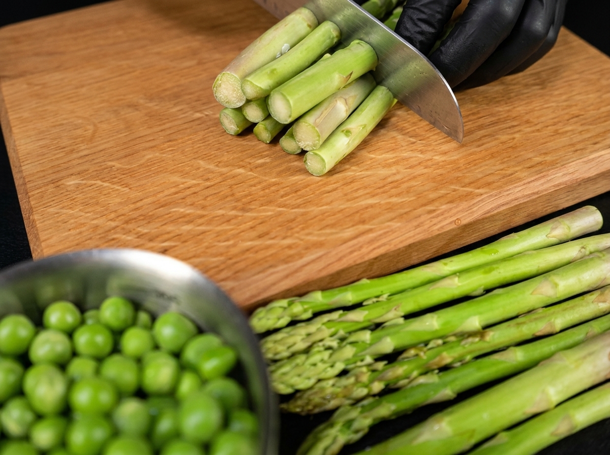 A chefs hand with a knife cutting fresh green asparagus stalks into even pieces on a wooden surface.