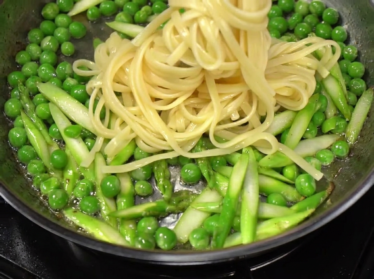 Spaghetti being lifted from boiling water and added to a pan of stir-fried vegetables and bacon.