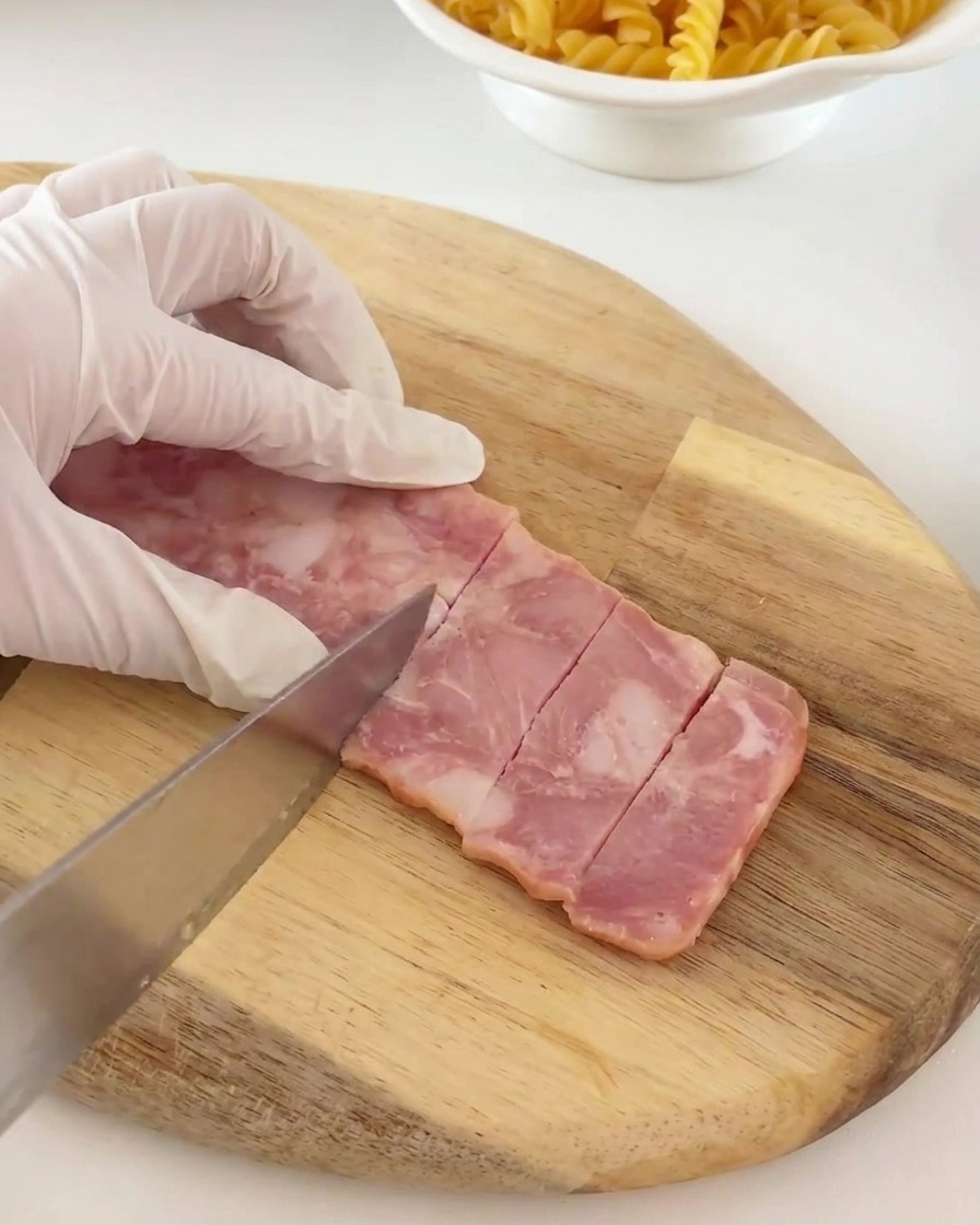 Strips of bacon being cut into small, consistent rectangular pieces on a wooden board.