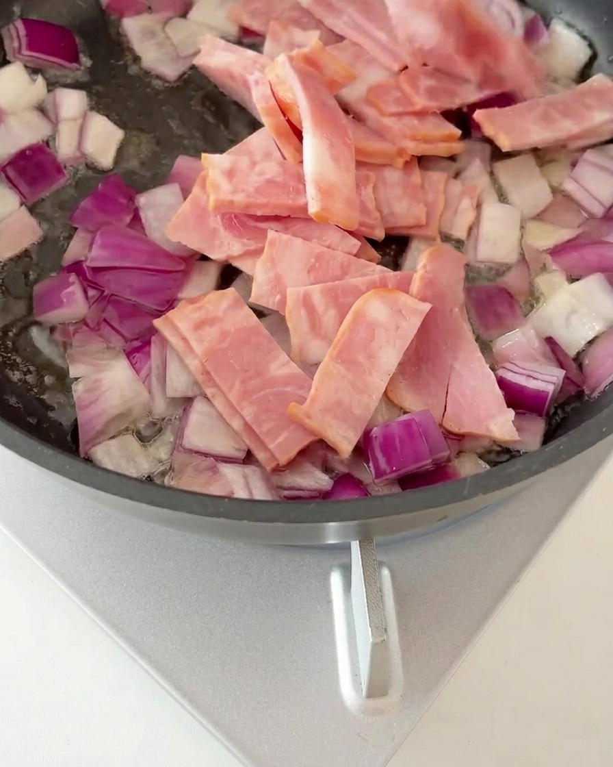 Bacon pieces mixed with sautéed onions in a frying pan, starting to brown.
