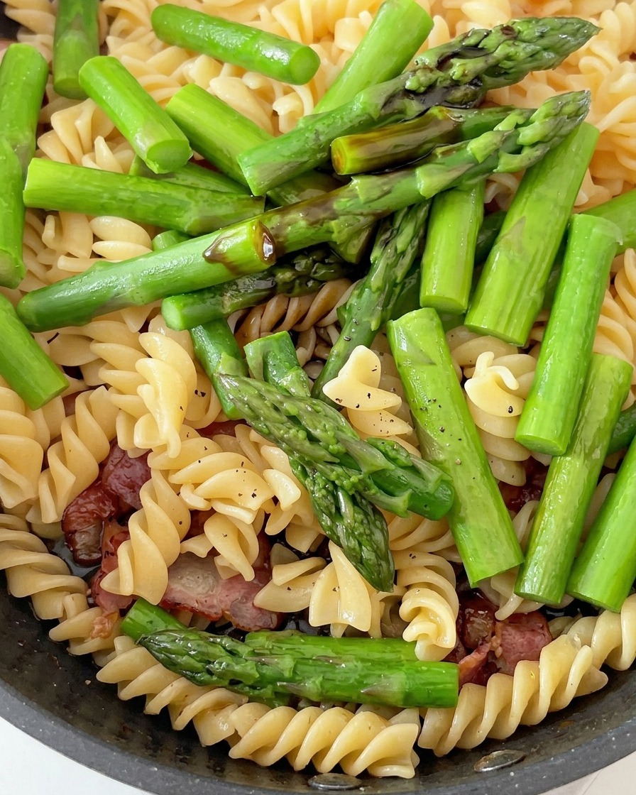 A pan containing cooked fusilli pasta, vibrant green asparagus pieces, and cooked bacon, being seasoned with sauces.