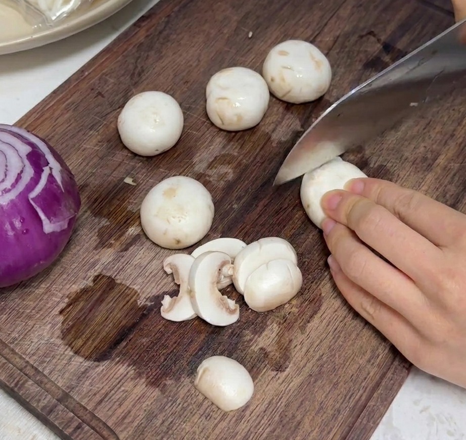 Fresh white mushrooms being sliced into even pieces on a wooden cutting board.