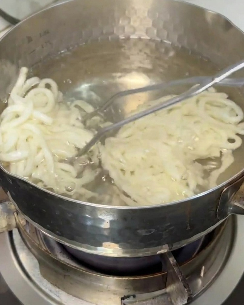 Udon noodles being lifted out of a pot of boiling water with a kitchen strainer.