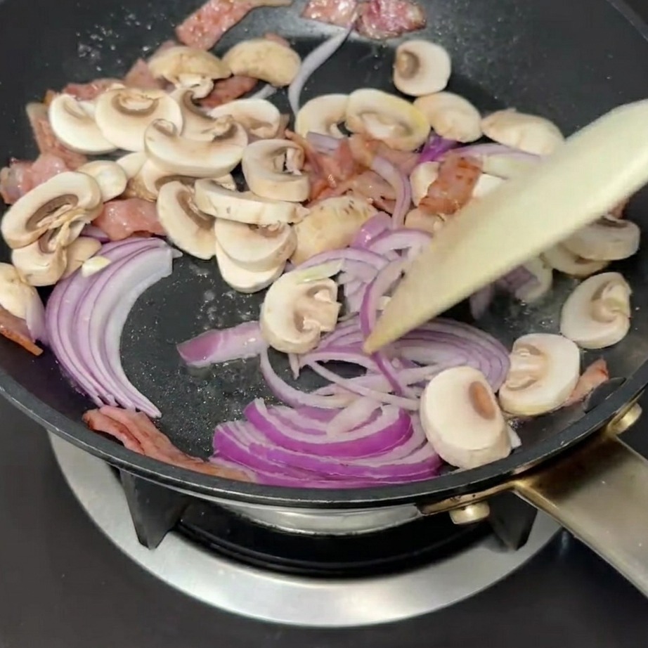 Sliced red onions and white mushrooms being stirred in a pan with bacon.