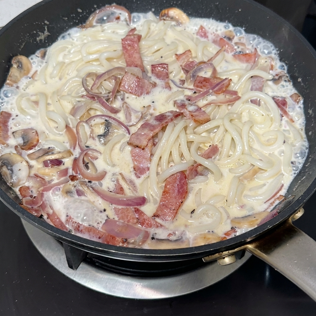 Black truffle paste being stirred into a pan filled with crispy bacon, sliced mushrooms, and caramelized onions.