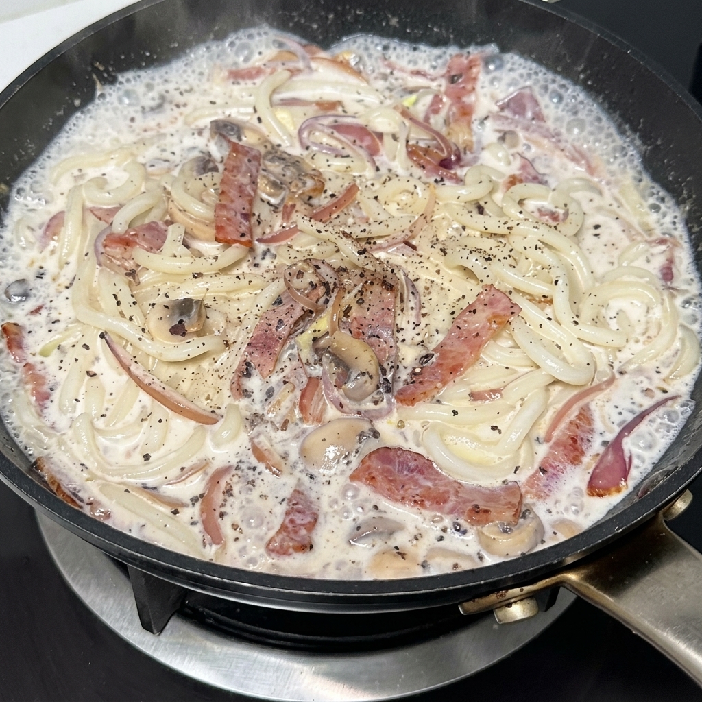 A pan of creamy udon noodles with bacon and mushrooms being seasoned with black pepper.