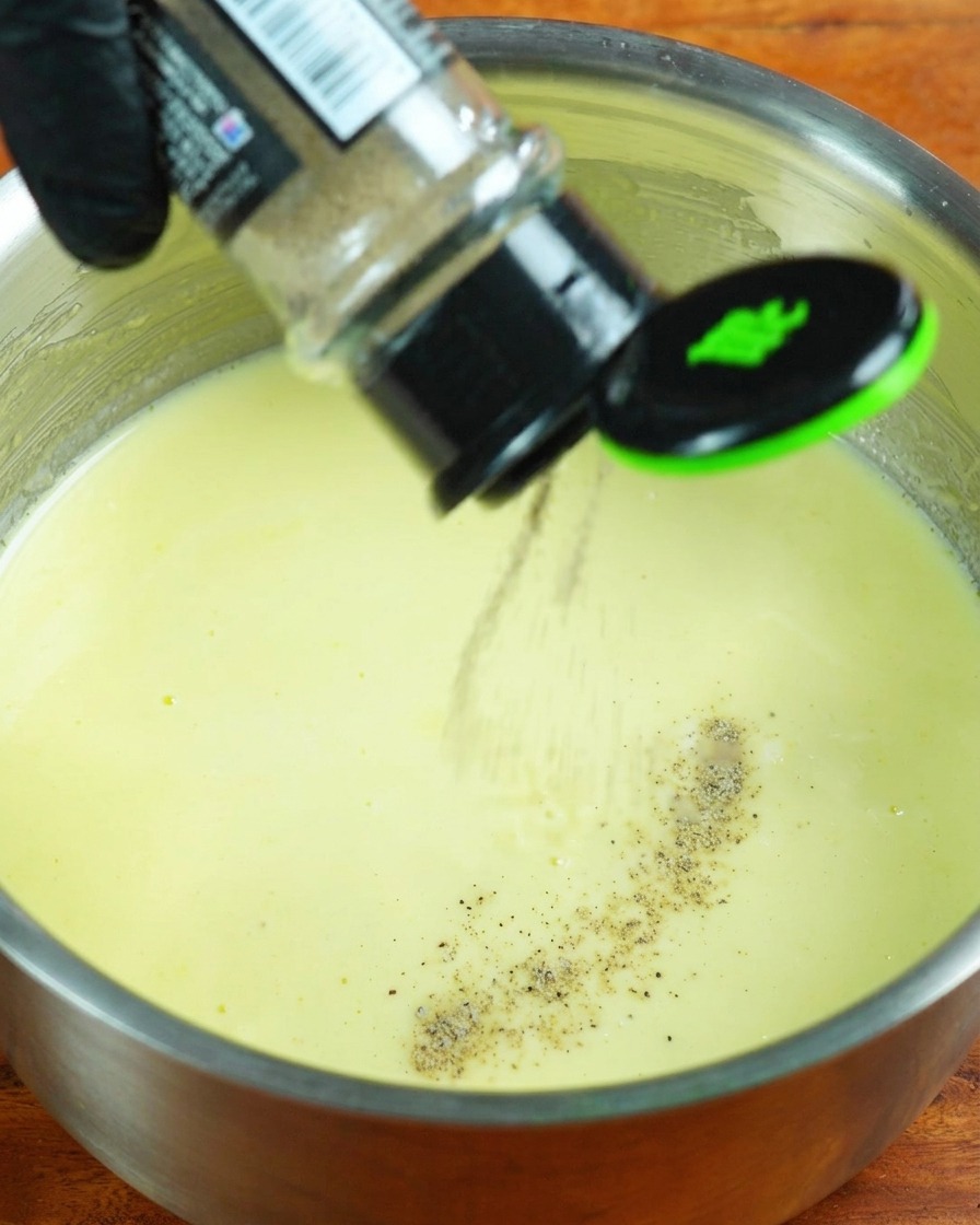 Heavy cream, white pepper, and salt being added to the simmering milk sauce.