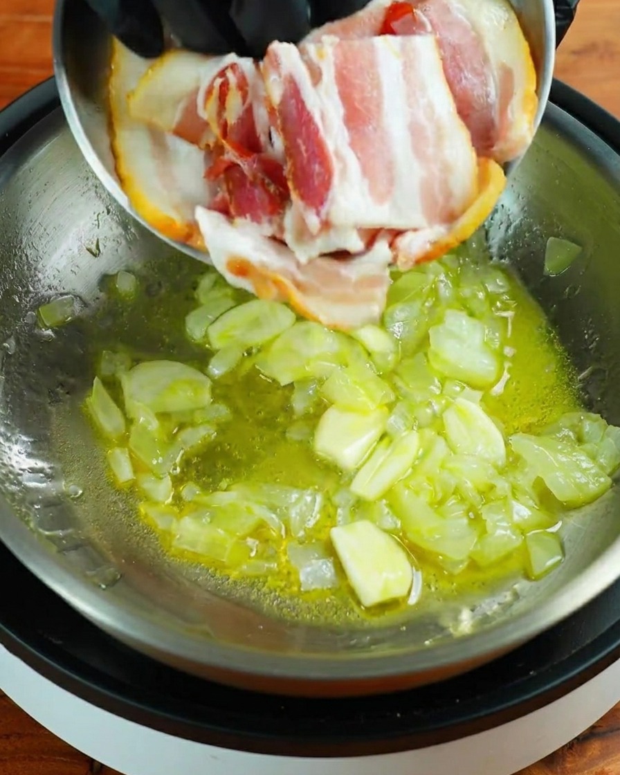 Three strips of raw bacon being added to a stainless steel pan containing sautéed garlic and onion pieces.