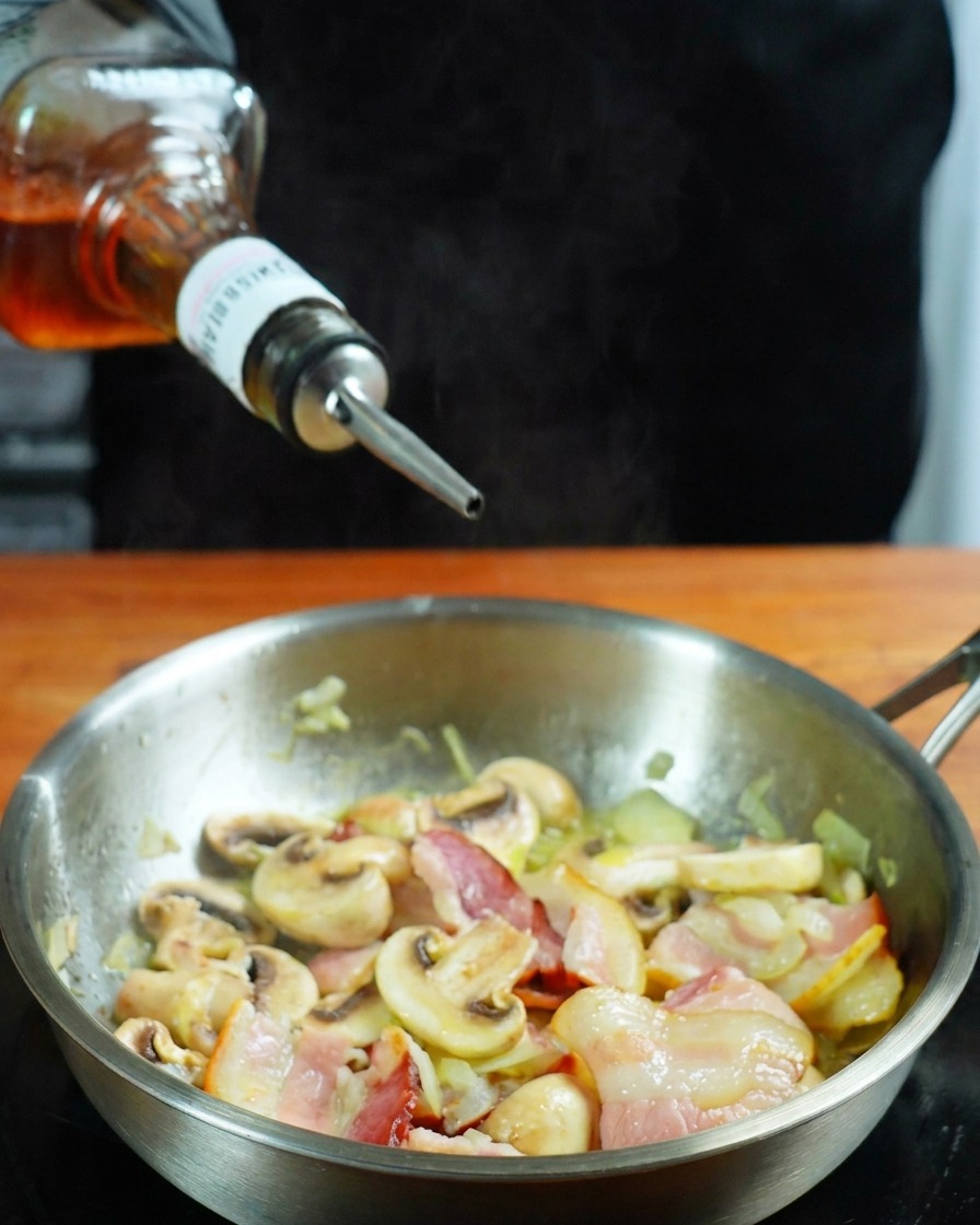 Whiskey being poured from a bottle into a stainless steel pan containing bacon and mushrooms.