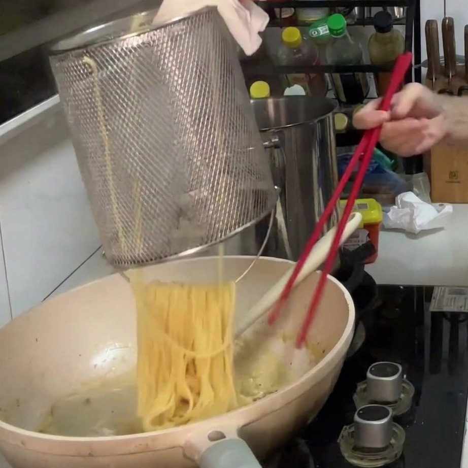 Boiled spaghetti being transferred from a metal basket into a pan with creamy mushroom sauce.
