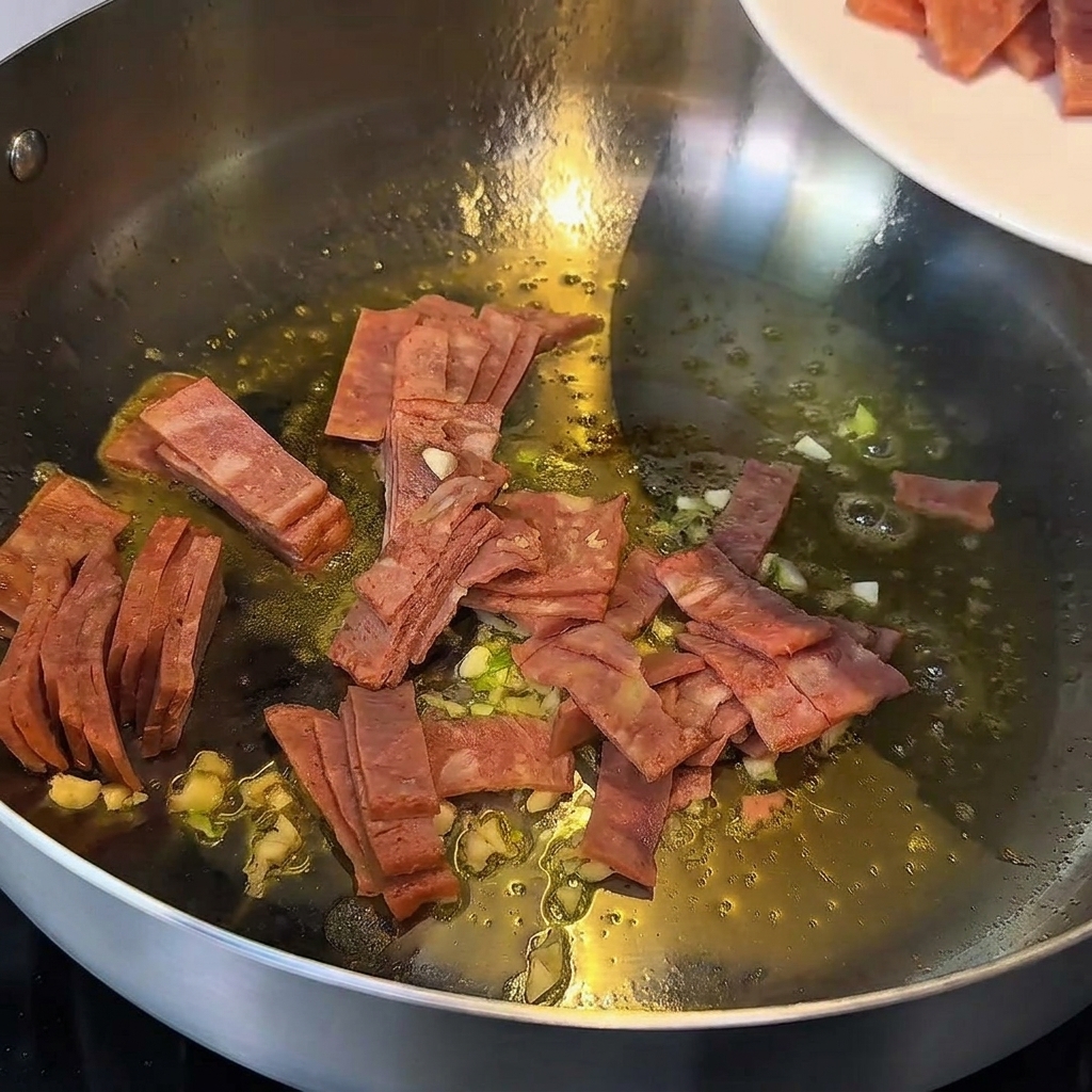 Garlic and bacon pieces being stir-fried in melted butter in a metal pan.