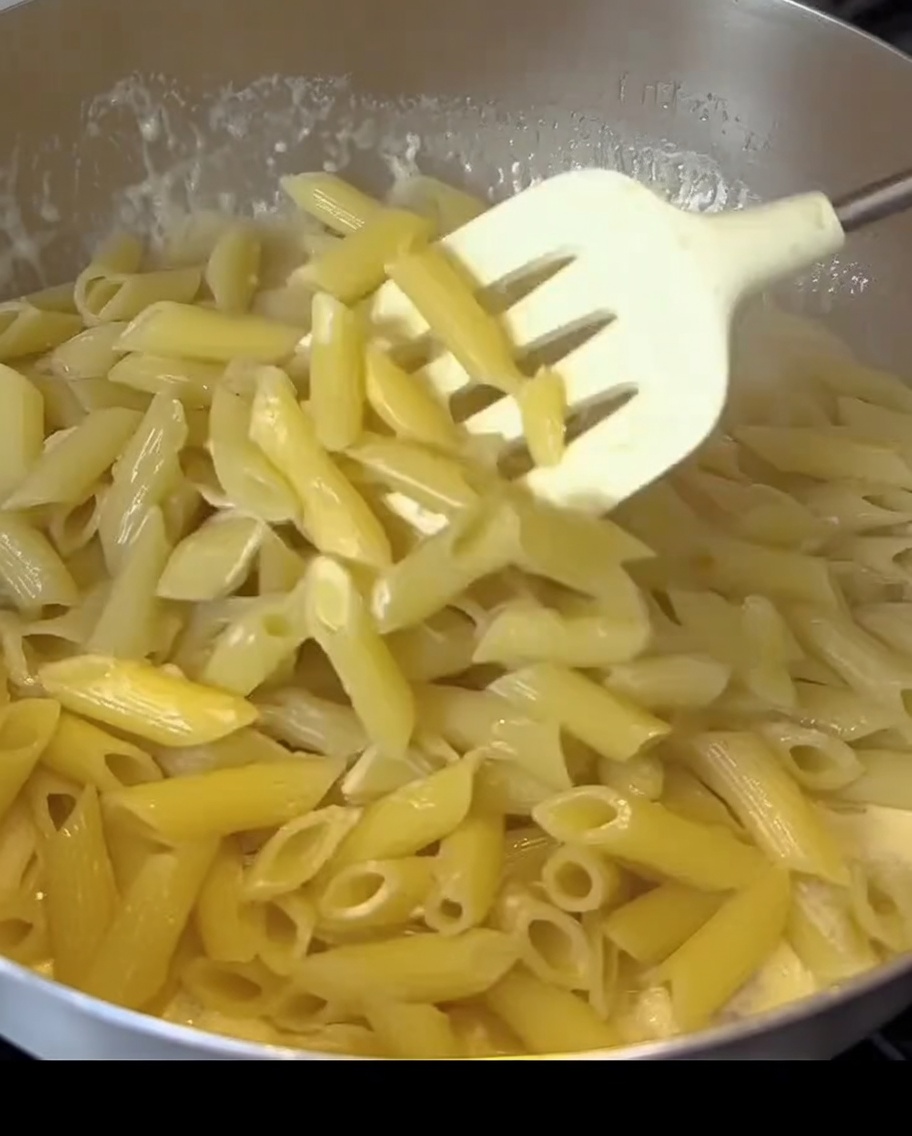 Penne pasta being stirred into a creamy white sauce with bacon pieces in a pan.
