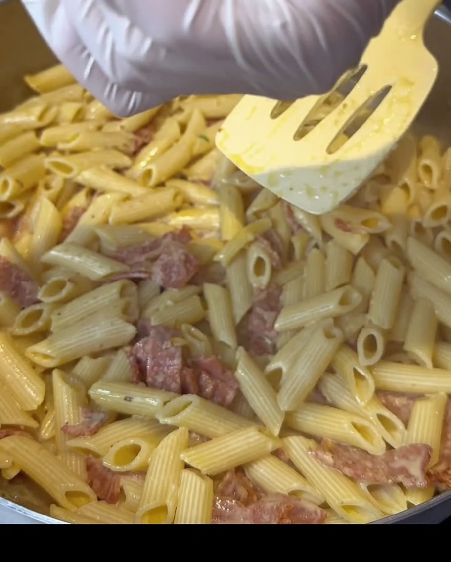 Gloved hands sprinkling seasoning over a pan of creamy bacon pasta.