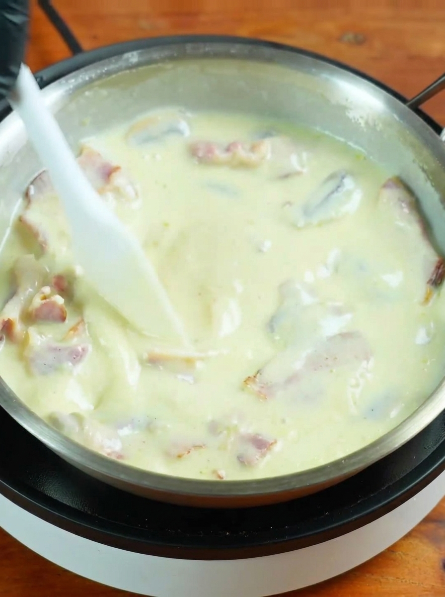 Cooked spaghetti being added to a bubbling white cream sauce in a large frying pan.