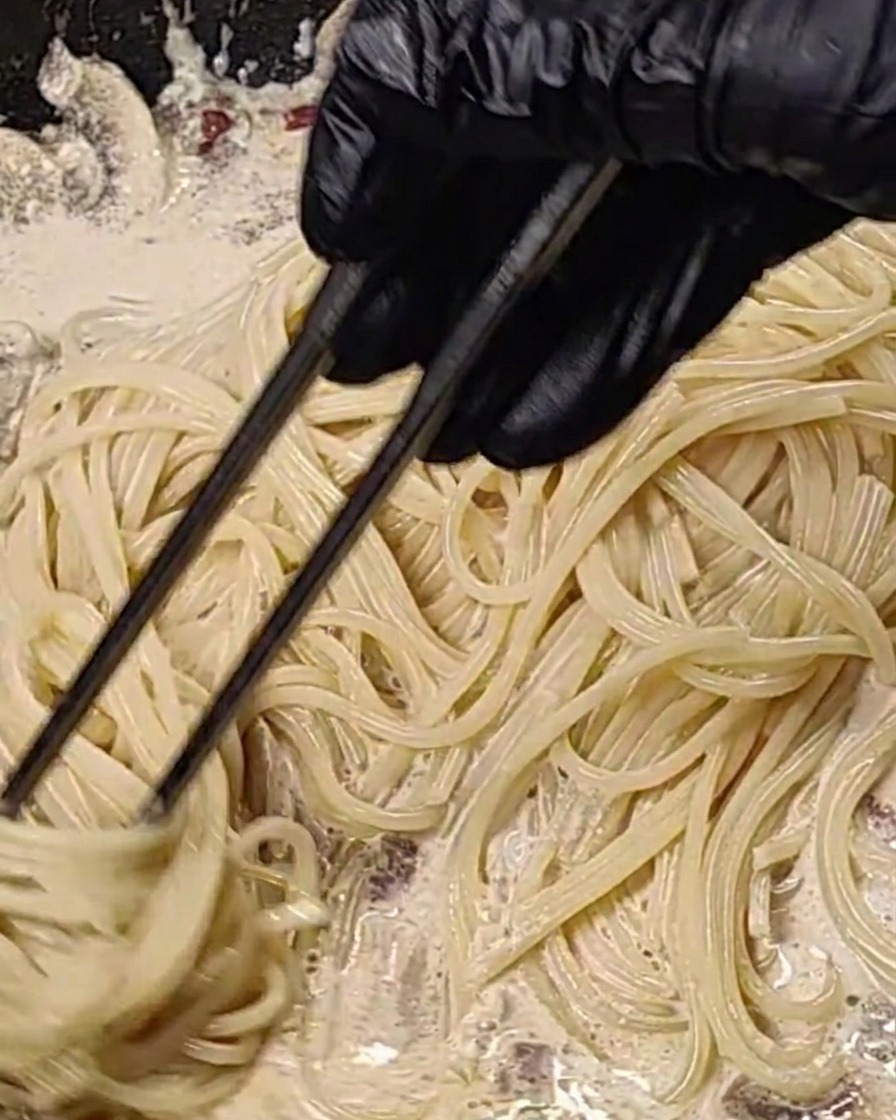Spaghetti being tossed in a creamy bacon and mushroom sauce in a hot pan.