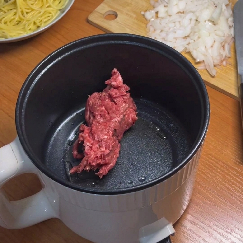 Minced beef being browned in a small electric pot with a light blue silicone spatula.
