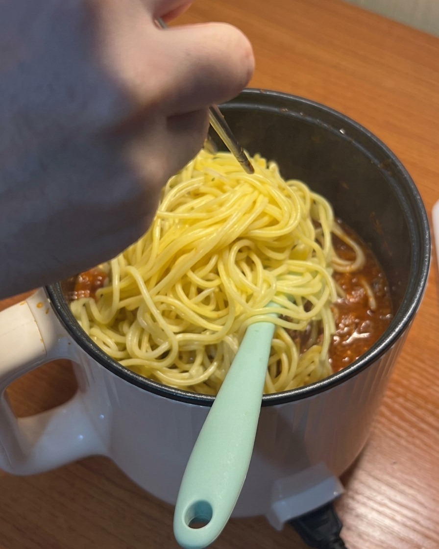 Cooked spaghetti being added to the simmering tomato meat sauce.