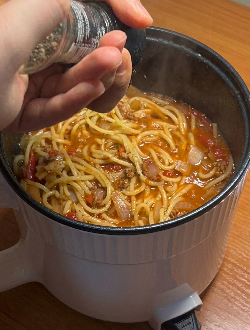 A close-up of a pot of finished spaghetti with meat sauce, seasoned with a fresh grating of black pepper.