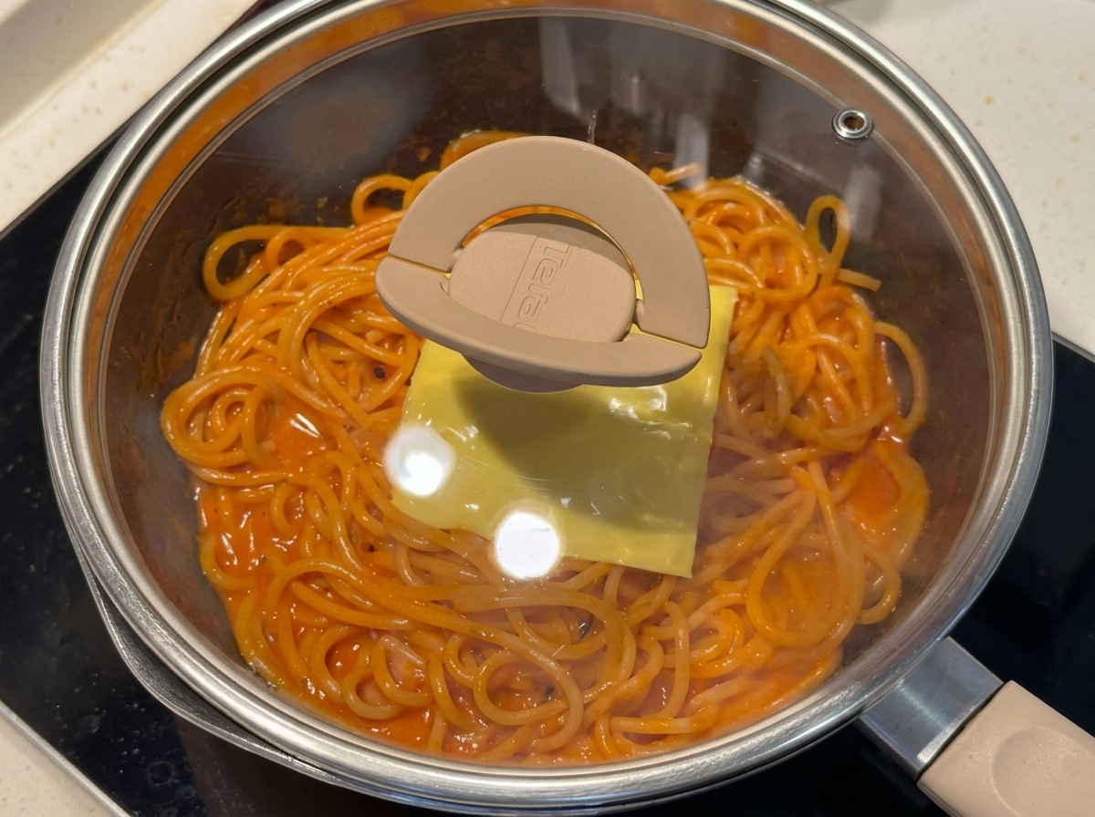 A pan of spaghetti topped with a slice of cheese covered by a glass lid.