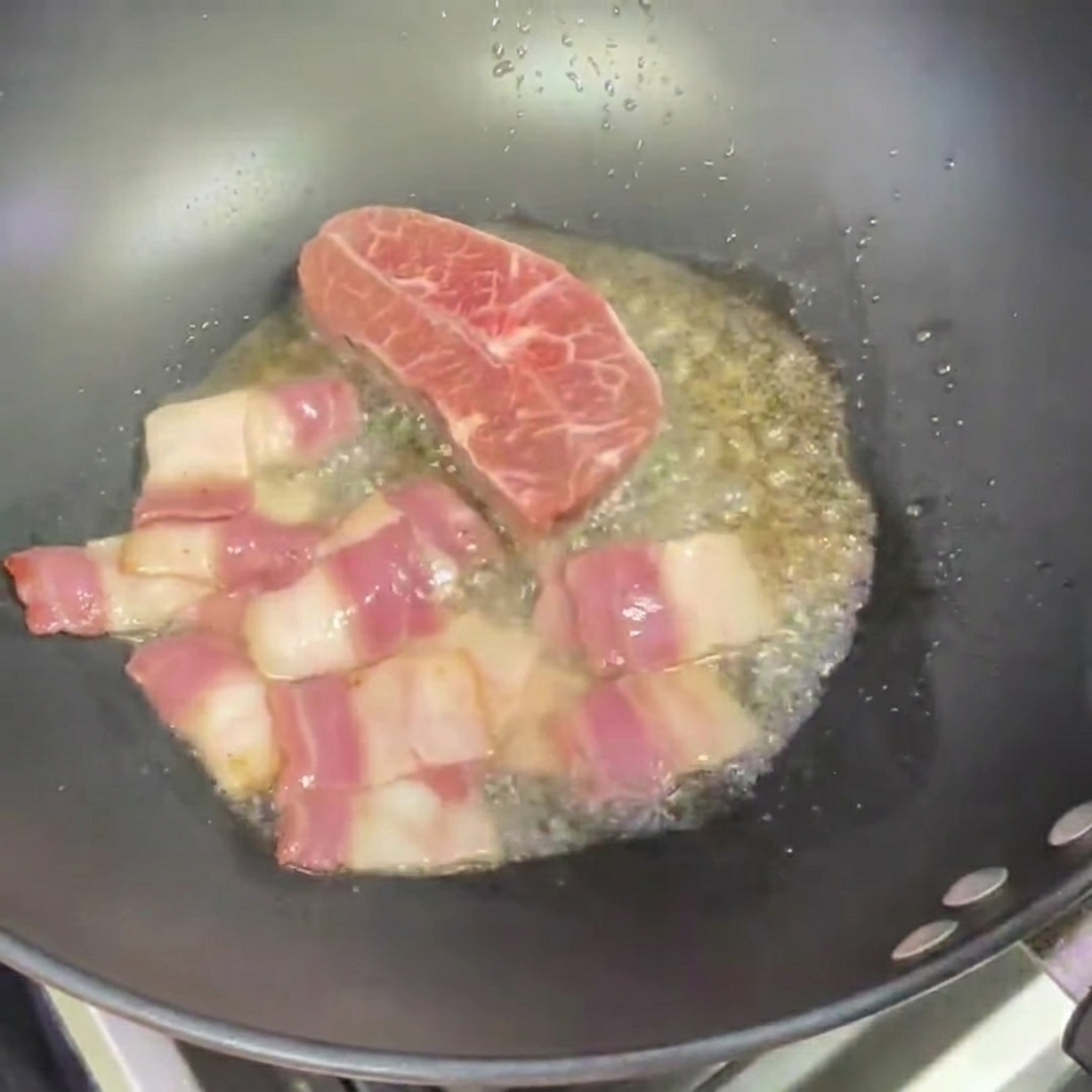 Two small steaks frying in a wok next to pieces of bacon.