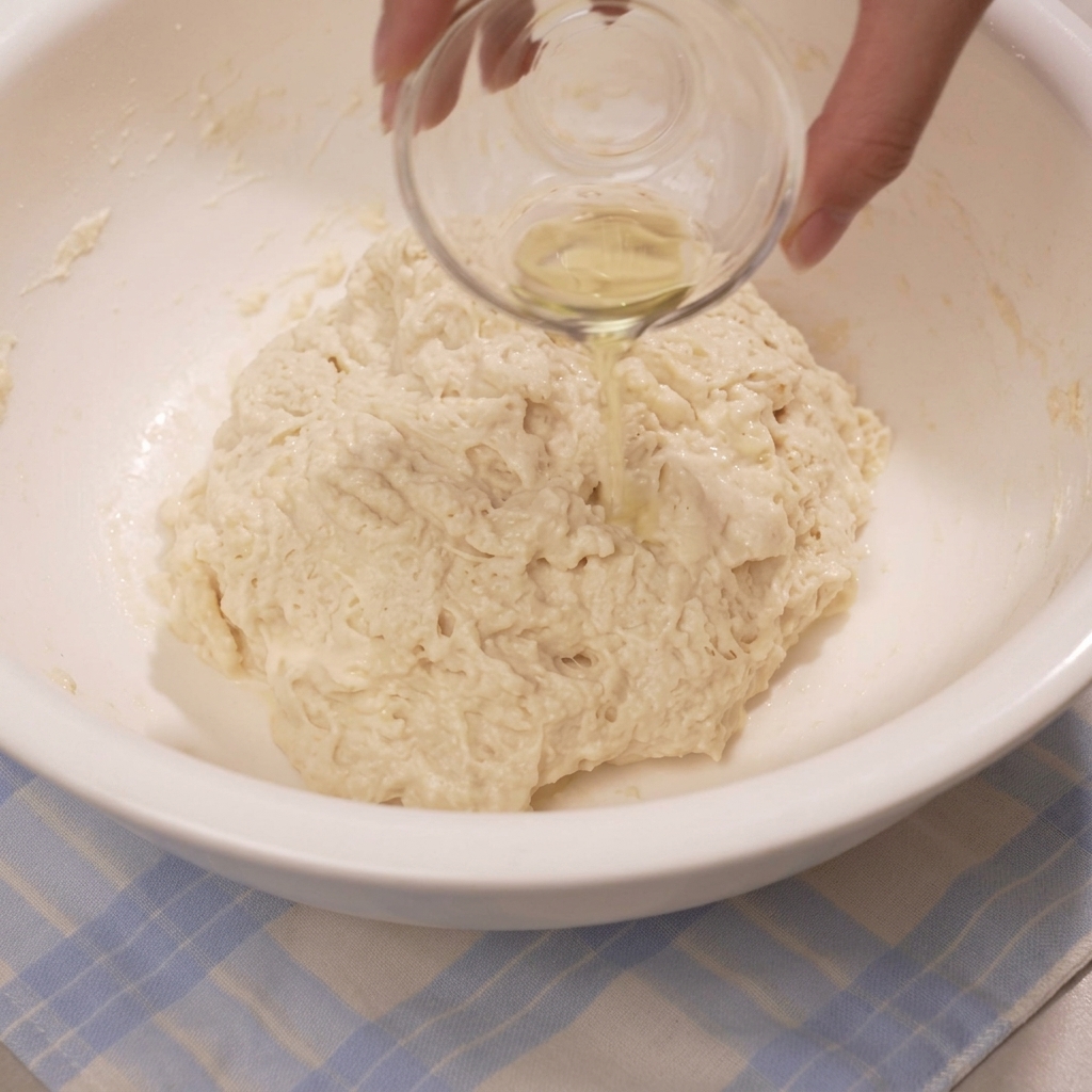 A hand pouring oil from a small glass bowl over a rough, shaggy dough mixture inside a large bowl.