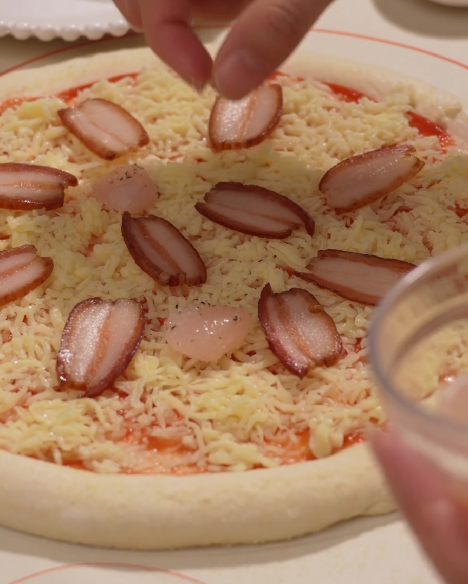 Ham slices and shrimp being carefully placed onto a cheese-topped pizza base.
